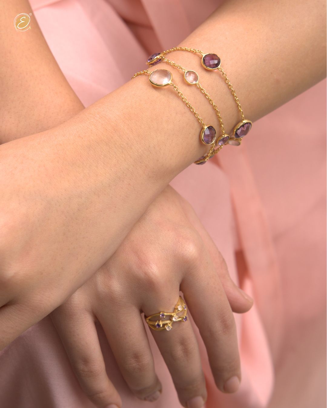 Gold bracelet and ring with gemstones on a wrist against a pink background